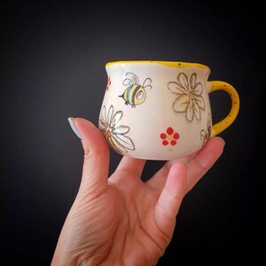 Puede incluir: Una pequeña taza de cerámica pintada a mano con asa y borde amarillos. La taza blanquecina presenta ilustraciones caprichosas de abejas, margaritas y racimos de flores rojas. La taza se sostiene en una mano sobre un fondo negro.