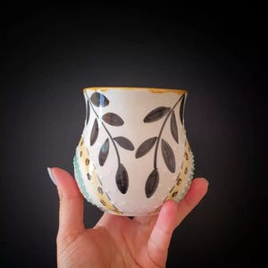 May include: A white ceramic vase with a black leaf design. The vase has a yellow rim and a textured surface with small, raised dots. The vase is held in a hand against a black background.