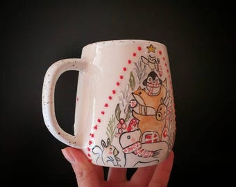 May include: A white ceramic mug with a whimsical winter scene featuring a penguin, fox, deer, and other animals. The mug has a speckled handle and is decorated with red dots and festive greenery. The design is hand-painted with a charming, illustrative style.