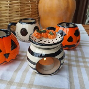 Puede incluir: Un quemador de aceite de cerámica con temática de Halloween con una vela encendida, rodeado de tazas con forma de calabaza y calabazas decorativas. El quemador de aceite tiene rayas blancas y negras, y las tazas son naranjas y blancas con detalles negros.