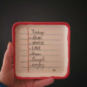May include: A small, rectangular ceramic dish with a red border. The dish features a handwritten list with the words "Today: -live -smile -LOVE -dream -laugh -enjoy" and a small smiley face.