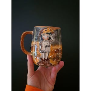 Puede incluir: Una taza de cerámica con una ilustración caprichosa de un niño con un sombrero de seta. La taza tiene un asa marrón y un diseño texturizado y terroso con una paleta de colores marrón, beige y gris. El niño lleva un vestido y botas de color marrón claro.