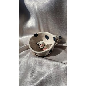 May include: A white ceramic mug with black polka dots and a small handle. Inside the mug is a miniature ceramic pig with black spots and pink accents. The mug is set against a silver fabric background.