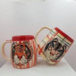Op de afbeelding: Twee keramische mokken met tijgergezichtontwerpen. Eén mok heeft een rode rand en verticale rood-witte strepen, terwijl de andere een ruitpatroon heeft. Beide mokken hebben een tijgergezicht met zwarte en oranje details.