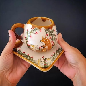 Peut inclure: Un ensemble tasse et soucoupe en céramique peinte à la main. La tasse et la soucoupe sont blanches avec un bord et une anse marron. L'ensemble présente une scène de forêt avec un renard et des lapins, entourés de feuilles vertes et de baies rouges.