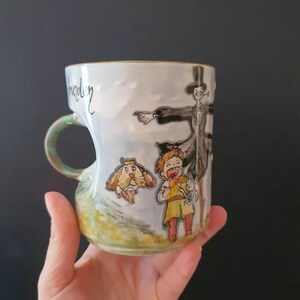May include: A ceramic mug with a whimsical illustration featuring a cartoon character, a dog, and a scarecrow. The mug has a green handle and a light blue and white background. The word "burden" is written on the side.