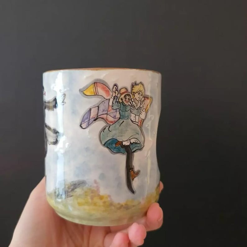 May include: A white ceramic mug with a hand-painted illustration of two figures in a whimsical style. The figures are depicted with a colorful blanket and a light blue background. The mug has a gold rim.