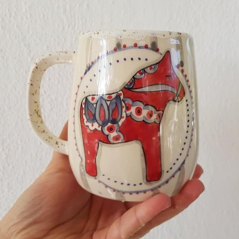 Może przedstawiać: Kremowy ceramiczny kubek z czerwono-niebieskim wzorem konia Dala. Kubek ma nakrapiany uchwyt i ozdobny brzeg. Koń znajduje się w kropkowanym okręgu. Unikalny element do kawy lub herbaty.