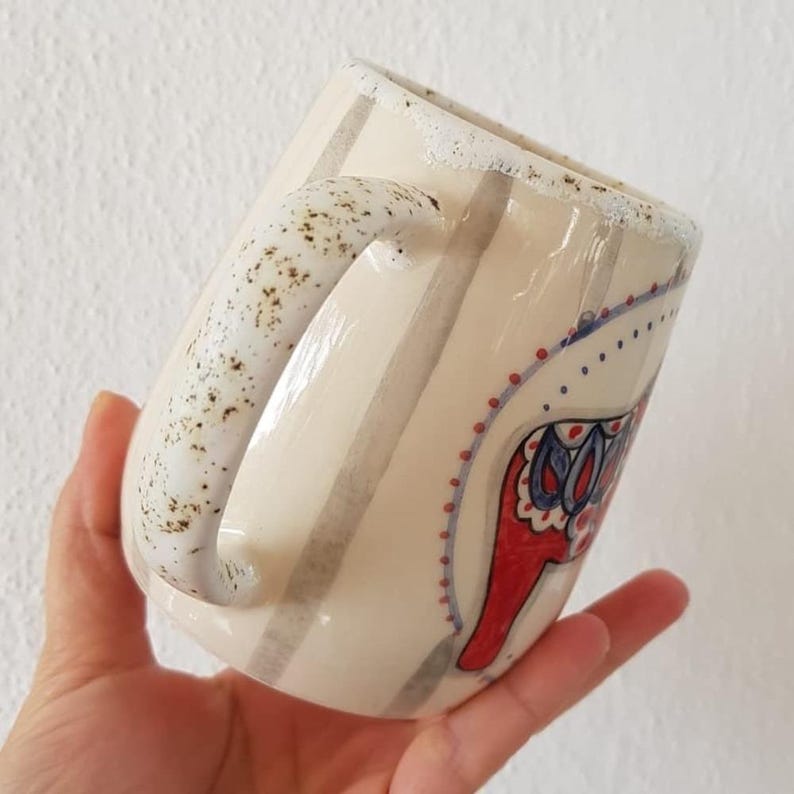 Może przedstawiać: Kremowy ceramiczny kubek z pionowymi szarymi paskami i nakrapianym uchwytem. Kubek ma czerwono-niebieski kwiatowy wz&oacute;r. Obręcz ma teksturowane, białe wykończenie. Unikalny, ręcznie robiony element do gorących napoj&oacute;w.