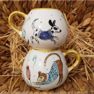 May include: Two stacked, white ceramic mugs with yellow handles and rims. The top mug features a running dog illustration wearing a blue sweater. The bottom mug has a stretching cat illustration, also in a blue sweater. Floral and bone accents decorate the mugs.