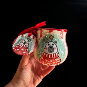 May include: A ceramic Christmas mug with a polar bear design. The mug is white with red stripes and a red handle. The polar bear is wearing a red sweater with white hearts and a star on its head. A matching tag is attached with a red ribbon.