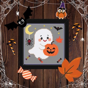 May include: A framed cross-stitch featuring a smiling ghost holding a jack-o'-lantern. The design includes a crescent moon, stars, a spider, and a maple leaf. Halloween-themed decorations, including candy and a bat, surround the frame.