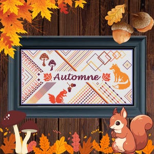 May include: Cross-stitch artwork in a black frame with the word "Automne" and autumn-themed imagery. The design includes a fox, squirrel, mushrooms, and geometric patterns in orange, brown, and white. The background is a wooden surface.