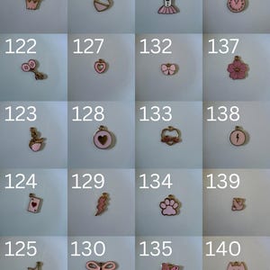 K&ouml;nnte beinhalten: Eine Sammlung von zwanzig kleinen Metallanh&auml;ngern mit rosa Emaille-Details, die jeweils mit einer Nummer von 121 bis 140 beschriftet sind. Die Anh&auml;nger haben verschiedene Formen, darunter eine Krone, Herzen, ein Kleid, eine Uhr, ein Schwan, eine Spielkarte, ein Stern, eine Schleife, eine Pfote, ein Schmetterling, ein Blitz, eine Blume und ein Katzengesicht.