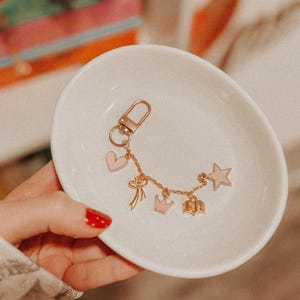 K&ouml;nnte beinhalten: Ein goldfarbener Metallschl&uuml;sselanh&auml;nger mit Karabinerverschluss und einer feinen Kette, die mit f&uuml;nf Anh&auml;ngern verziert ist: ein hellrosa Herz, eine Schleife, eine hellrosa Krone, ein offenes Buch und ein hellrosa Stern, in einer kleinen wei&szlig;en Keramikschale.