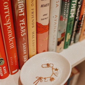 May include: A small, round, white ceramic trinket dish holding a gold-toned charm bracelet. The bracelet features several delicate charms, including a bow, a heart, a small building, a flower, and a star, all attached to a thin gold chain.