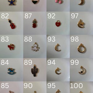 K&ouml;nnte beinhalten: Eine Sammlung von zwanzig kleinen Metallanh&auml;ngern, die in einem Raster angeordnet und von 81 bis 100 nummeriert sind. Die Anh&auml;nger haben verschiedene Formen, darunter ein blauer Seestern, eine rote Gitarre, eine Katze, ein Schaukelpferd, ein Weinglas, ein Herz, eine Ananas, ein Oktopus, ein Kaktus, ein Stern und Monde.