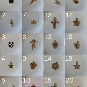 K&ouml;nnte beinhalten: Eine Sammlung von zwanzig kleinen Metallanh&auml;ngern in einem Raster, darunter Formen wie Kaffeetasse, Blitz, Schachbrett-Herz, Sterne, Schwert, Schleife, Kreuz, K&uuml;rbis und Mond, teilweise mit Strasssteinen, Emaille oder Perlen.