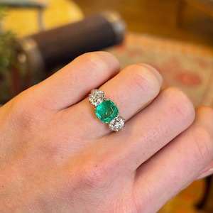 Könnte beinhalten: Ein Ring mit einem großen, quadratisch geschliffenen Smaragd-Mittelstein, flankiert von zwei Clustern kleinerer, runder, klarer Edelsteine. Der Ring ist in einem hellen Metall, wahrscheinlich Gold oder Platin, gefasst.