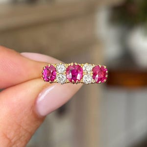 Puede incluir: Un anillo de oro con cinco piedras preciosas rosas ovaladas y vibrantes, alternando con cuatro piedras preciosas claras, redondas y más pequeñas. El engaste del anillo es de color oro brillante.