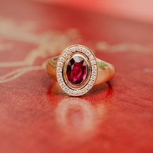May include: A gold ring featuring a central oval-cut deep red ruby gemstone. The ruby is surrounded by a halo of small, clear, round-cut white diamonds set within a gold frame. The band of the ring is smooth, polished gold with a tapered design.