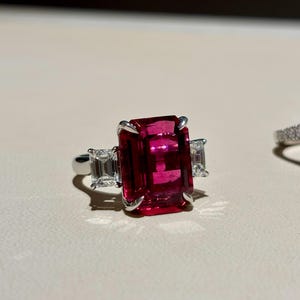 Könnte beinhalten: Ein silberfarbener Ring mit einem großen, rechteckigen, tiefrosa Edelstein in der Mitte. Der zentrale Stein wird von zwei kleineren, klaren, rechteckig geschliffenen Steinen flankiert, die alle von Metallfassungen auf einer glatten Ringschiene gehalten werden.