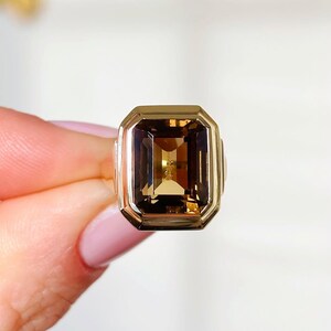 May include: A gold-toned ring featuring a large, rectangular, smoky-brown gemstone. The ring has a classic, geometric design with a polished finish. The gemstone is set within a gold bezel, creating a bold and elegant statement piece.