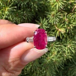 May include: A ruby ring with a large oval-cut, deep red gemstone. The ring has a silver band with two baguette-cut diamonds on either side of the main stone. The ring is set against a backdrop of green foliage.