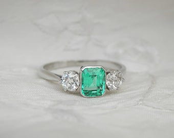 May include: A silver ring with a rectangular emerald centre stone and two round, clear gemstones. The emerald is a vivid green, and the other stones are clear. The ring is set against a white, textured background.