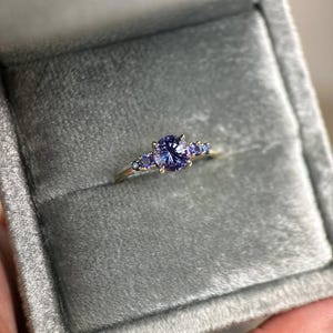 Op de afbeelding: Een gouden ring met een grote, ronde, paarse edelsteen in het midden, geflankeerd door kleinere paarse stenen. De ring wordt gepresenteerd in een grijze fluwelen sieradendoos.