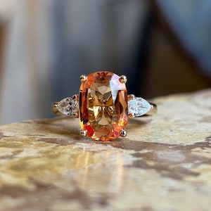 Peut inclure: Une bague en or ornée d'une grande pierre précieuse orange de forme ovale, flanquée de deux pierres blanches en forme de poire. La bague est posée sur une surface en marbre, mettant en valeur le design et la couleur du bijou.