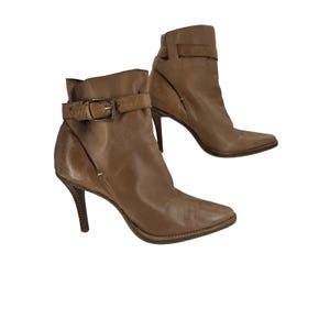 Könnte beinhalten: Ein Paar knöchelhohe Stiefel aus beigem Leder mit spitzer Zehenpartie, schmalem Stiletto-Absatz und einem dekorativen Riemen am Knöchel, der mit einer Metallschnalle befestigt ist. Die Stiefel haben eine glatte Oberfläche und eine strukturierte Form.