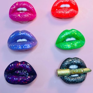 Puede incluir: Seis accesorios con forma de labios coloridos en rosa, rojo, azul, verde, morado y negro, cada uno con dientes blancos. El accesorio de labios negro tiene una bala dorada. Los accesorios están sobre un fondo rosa.