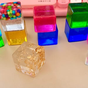 May include: A selection of colourful resin cubes. Some cubes are clear with embedded decorations like gold flakes, colourful sprinkles, and snowflakes. Other cubes are solid colours, including blue, green, pink, and yellow. The cubes are arranged on a light-coloured surface.
