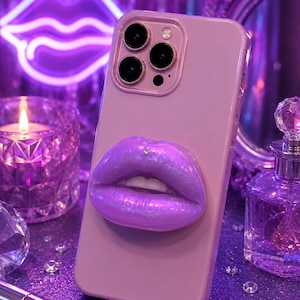 Puede incluir: Una funda de smartphone de color morado claro con un agarre tridimensional en forma de labios morados iridiscentes y brillantes en la parte posterior. Los labios tienen un pequeño brillante transparente centrado en el labio superior. Tiene recorte.