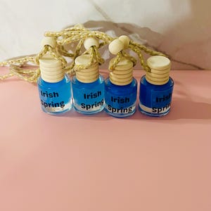 May include: Four small glass bottles filled with blue liquid, each with a wooden cap and a gold string for hanging. The bottles are labeled "Irish Spring" in white text. The bottles are arranged on a pink surface.