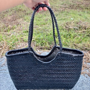 May include: A black woven tote bag with braided handles. The bag has a rectangular shape with a curved opening and a textured, basket-like weave. The handles are thick and sturdy, suitable for carrying various items.