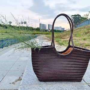 May include: A dark brown woven tote bag with a braided handle, filled with green foliage. The bag is sitting on a stone surface, with a blurred background of a park and buildings.