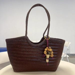 Puede incluir: Un bolso tote tejido marrón oscuro con un asa trenzada. El bolso tiene un adorno decorativo con varias cuentas y una borla morada. El bolso está hecho de un material marrón oscuro.