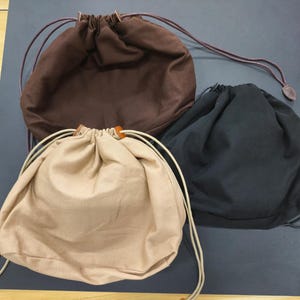 Könnte beinhalten: Drei Kordelzugbeutel in verschiedenen Farben: Braun, Beige und Schwarz. Jeder Beutel hat einen Kordelzugverschluss mit Lederakzent. Die Beutel bestehen aus einem weichen Stoff und eignen sich für verschiedene Zwecke.