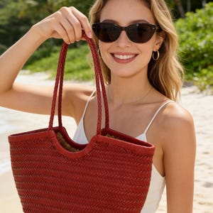Puede incluir: Un bolso tote rojo tejido con asas trenzadas. El bolso lo sostiene una persona que lleva gafas de sol y un vestido blanco. El bolso tiene forma rectangular y una textura tejida, perfecto para un día en la playa.