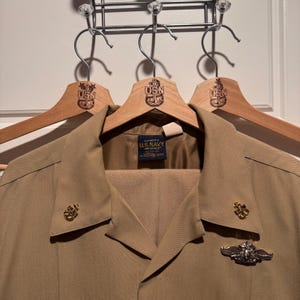 May include: A tan U.S. Navy uniform, including a jacket and trousers, hangs on a wooden hanger. The hanger is branded with the USN logo. The jacket features gold-colored insignia and a patch that reads "U.S. Navy Originals".