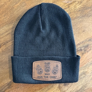 May include: A dark navy blue knitted beanie hat with a folded brim. A brown leather patch is sewn on the front, featuring three anchors, the letters "USN", the year "1893", and the text "ASK THE CHIEF".