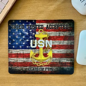 May include: A rectangular mouse mat with a distressed American flag design. The mat features the text "TRIED TESTED ACCEPTED", the letters "USN" with a gold anchor, and "CLASS 132 FY26". The design is set against a wooden background.