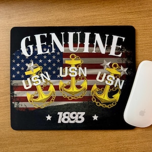 May include: Black mousepad with the word "GENUINE" in white, a distressed American flag design, and three gold anchors with the letters "USN". The number "1893" is below the anchors.