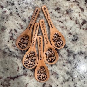 May include: Five wooden spoons with the text "Spoon of Knowledge" and a USN anchor design. The spoons are light brown and arranged on a speckled surface. The spoons vary slightly in size.