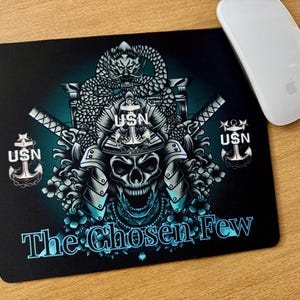 May include: Black mousepad with a skull design, samurai helmet, and the text "The Chosen Few" in teal. The design includes a dragon, crossed swords, and USN anchors. A white computer mouse is on the right.