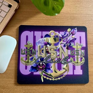 Peut inclure: Tapis de souris rectangulaire avec un fond violet et le mot "GIRL" en blanc. Le tapis de souris présente des ancres dorées, les lettres "USN" et des fleurs violettes. Le mot "Chick" est écrit en cursive. Une souris blanche et un ordinateur portable sont en arrière-plan.