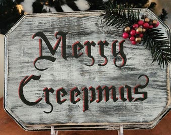 Puede incluir: Un letrero de madera rústico con las palabras "Merry Creepmas" en letras negras y rojas. El letrero tiene un fondo blanco y gris desgastado y está decorado con bayas artificiales y ramitas de pino, ideal para la decoración navideña.
