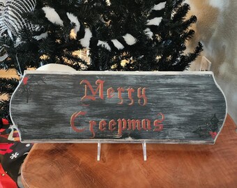 Puede incluir: Un letrero de madera gris oscuro con las palabras "Merry Creepmas" en letras rojas. El letrero tiene un acabado envejecido y está sostenido por un soporte transparente. Pequeñas rosas rojas y murciélagos decoran los bordes.